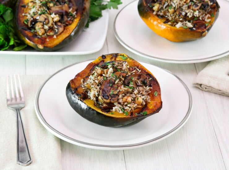 Vegan stuffed acorn squash filled with wild rice, mushrooms, and herbs on a white plate. Light wood table, and white tile background. A fork is placed on a beige napkin beside it.