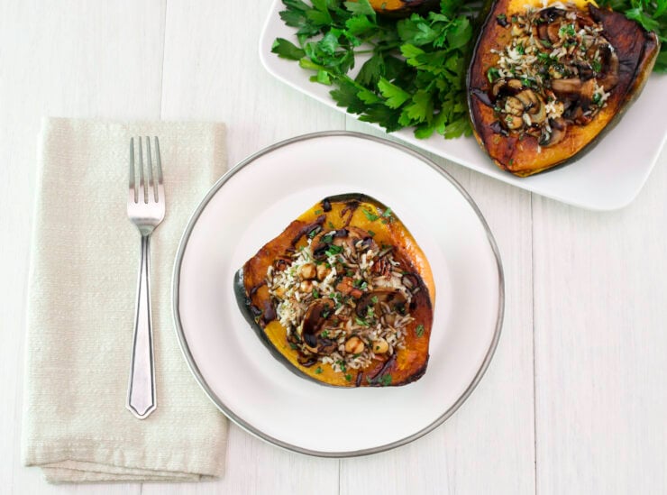 Vegan stuffed acorn squash is filled with rice, mushrooms, chickpeas, and herbs on a white plate. A fork is placed on a beige napkin beside it.
