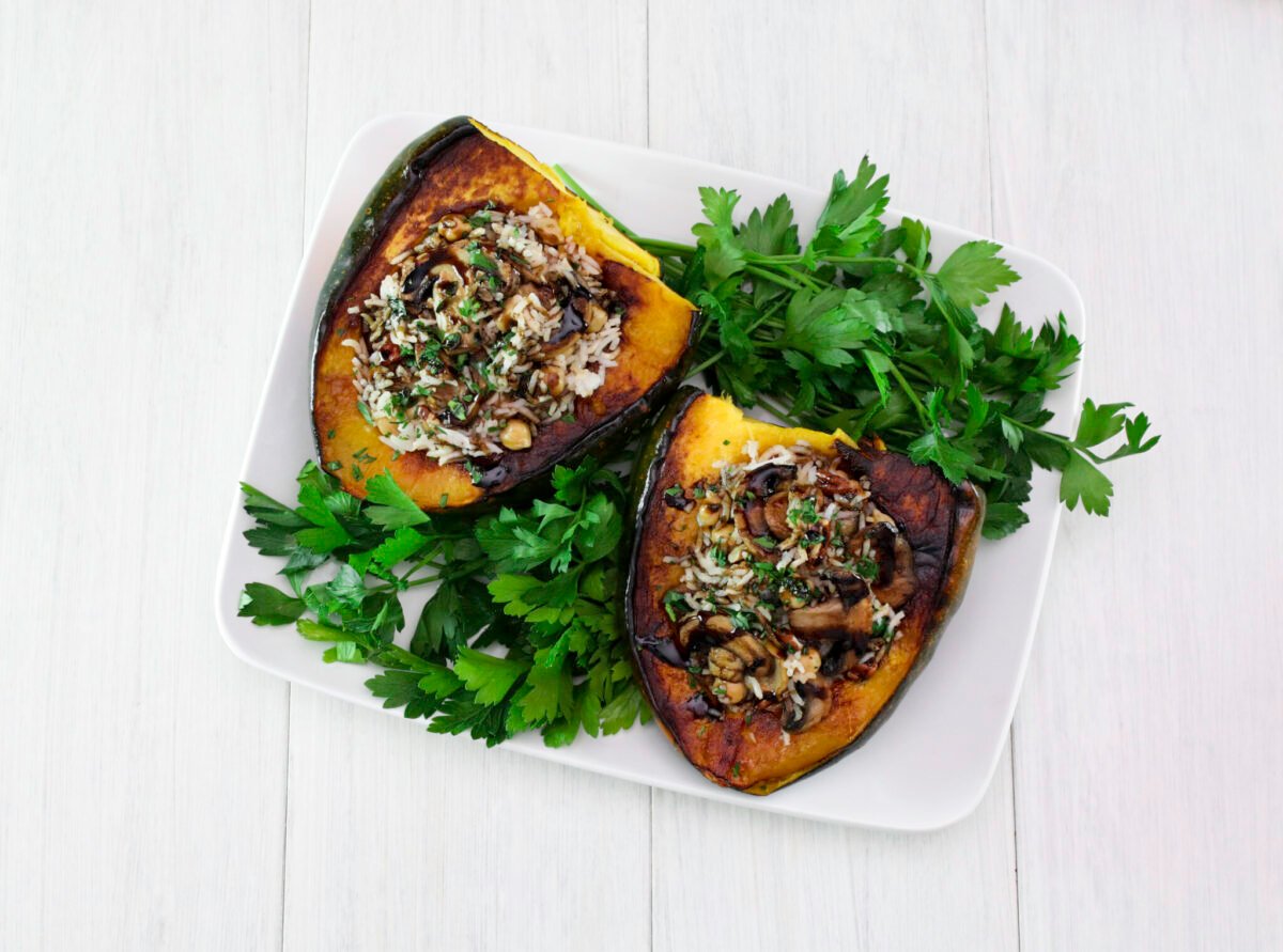 Two vegan stuffed acorn squash filled with rice, mushrooms, and herbs, garnished with fresh parsley on a white rectangular plate, on a light wooden table.Two vegan stuffed acorn squash filled with rice, mushrooms, and herbs, garnished with fresh parsley on a white rectangular plate, on a light wooden table.
