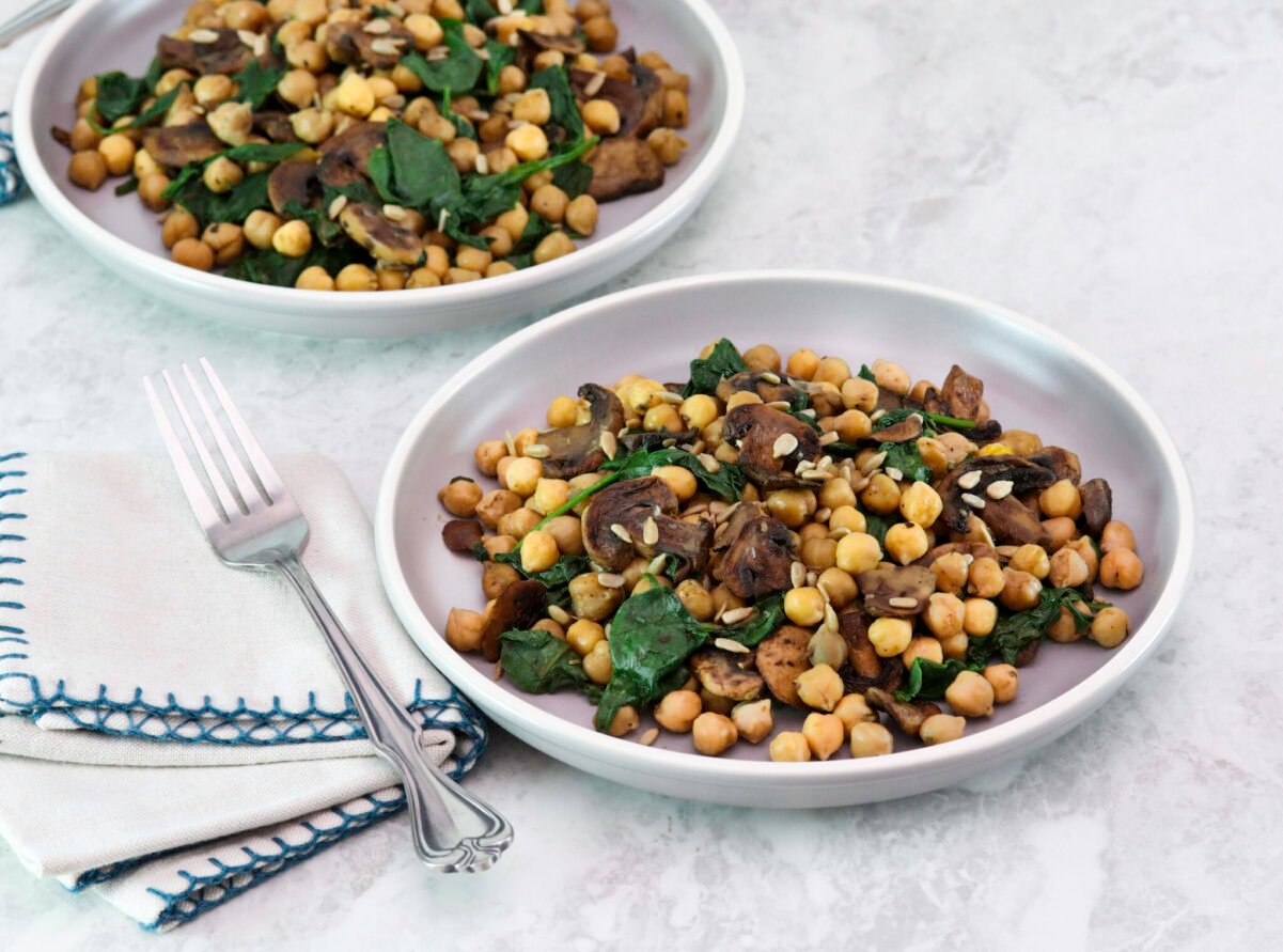 Two servings of sautéed chickpeas with spinach mushrooms on a white plate. A fork and linen napkin on the side.
