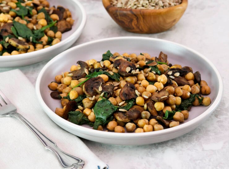 Two servings of sautéed chickpeas with spinach mushrooms on a white plate. A fork, linen napkin and a wooden bowl of seeds in the background.