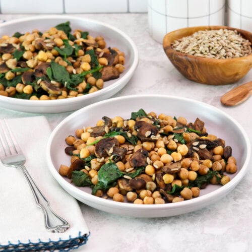 Two servings of sautéed chickpeas with spinach mushrooms on a white plate. A pair of fork, linen napkin, white canister, a wooden bowl of seeds and wooden spoon in the background.