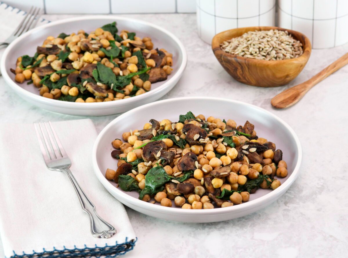 Two servings of sautéed chickpeas with spinach mushrooms on a white plate. A pair of fork, linen napkin, white canister, a wooden bowl of seeds and wooden spoon in the background.