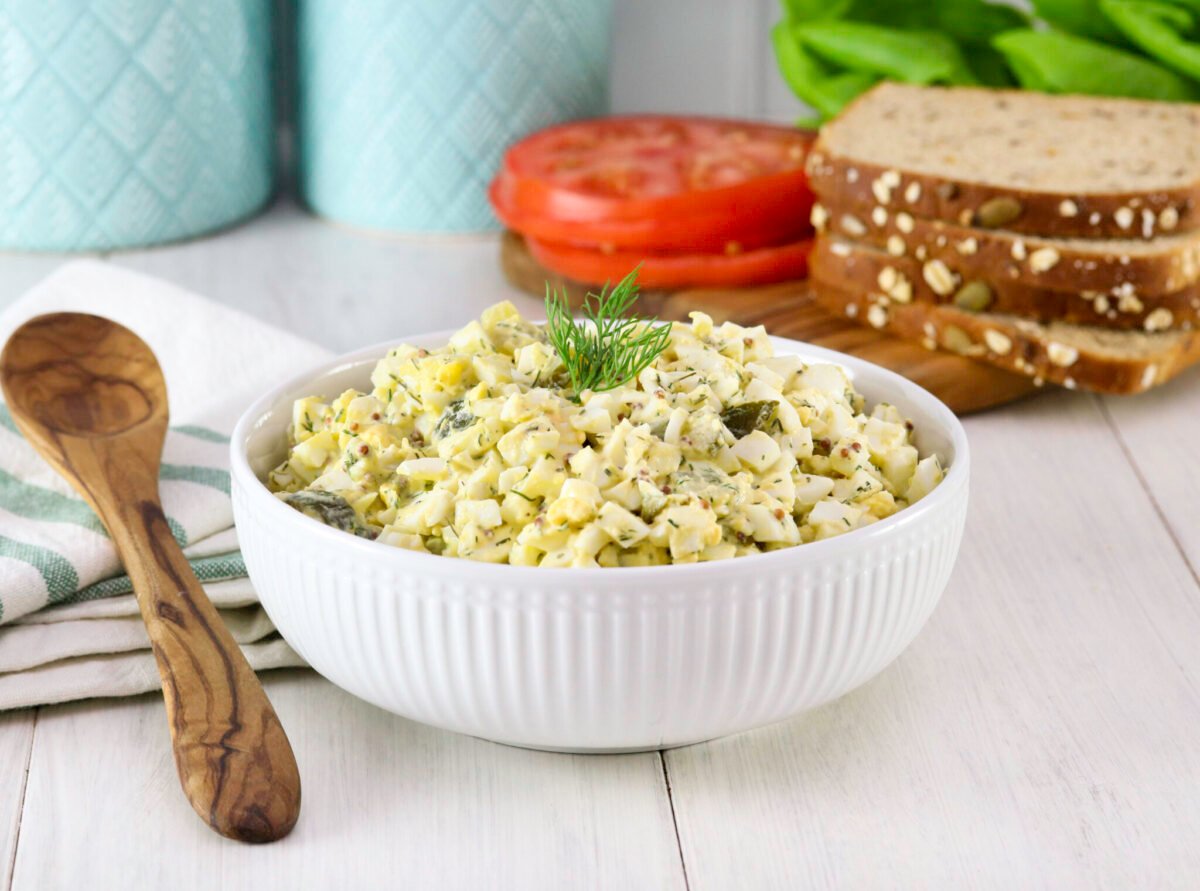 A bowl of egg salad garnished with dill sits on a wooden table. Nearby are sliced tomatoes, lettuce, seeded bread, turquoise jars, and a wooden spoon over a striped napkin.