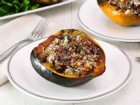Vegan Stuffed Acorn Squash Pinterest Pin - vegan stuffed acorn squash filled with wild rice, mushrooms, and herbs on a white plate. Light wood table, and white tile background. A fork is placed on a beige napkin beside it.