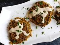 Kartoffelpuffer Pinterest Pin - Crispy baked potato tahini pancake patties topped with tahini and parsley on white plate with bowl of tahini, spoon, and cabbage salad in background, on grey concrete counter.