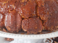 Monkey Bread Pin - Golden monkey bread with a sticky glaze sits on a white cake stand. Pinecones and a silver ribbon adorn the base, creating a festive atmosphere.